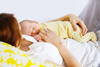 Mutter liegt in Schlafkleidung mit schlafendem Neugeborenen auf der Brust im Bett und hält sein Händchen