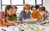Fröhliche Grundschulkinder stehen um einen Tisch herum und machen kreative Gruppenarbeit mit Legosteinen und schauen auf ein Tablet