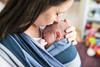 Mutter trägt schlafendes Neugeborenes im Wickeltuch an der Brust, hält mit einer Hand das Köpfchen und küsst dem Baby auf die Stirn