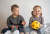 Ein Kleinkind mit Down-Syndrom und Stofftier und ein Kind ohne Beeinträchtigungen und mit gelbem Fußball in der Hand sitzen auf einer Decke und lachen 