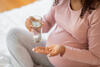 Hochschwangere sitzt im Schneidersitz auf dem Bett und hält in einer Hand ein Glas Wasser und in der anderen eine Tablette