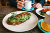 Toast mit Avocado im Vordergrund auf einem Tisch, an dem im Hintergrund eine Person mit einer Tasse Kaffee sitzt